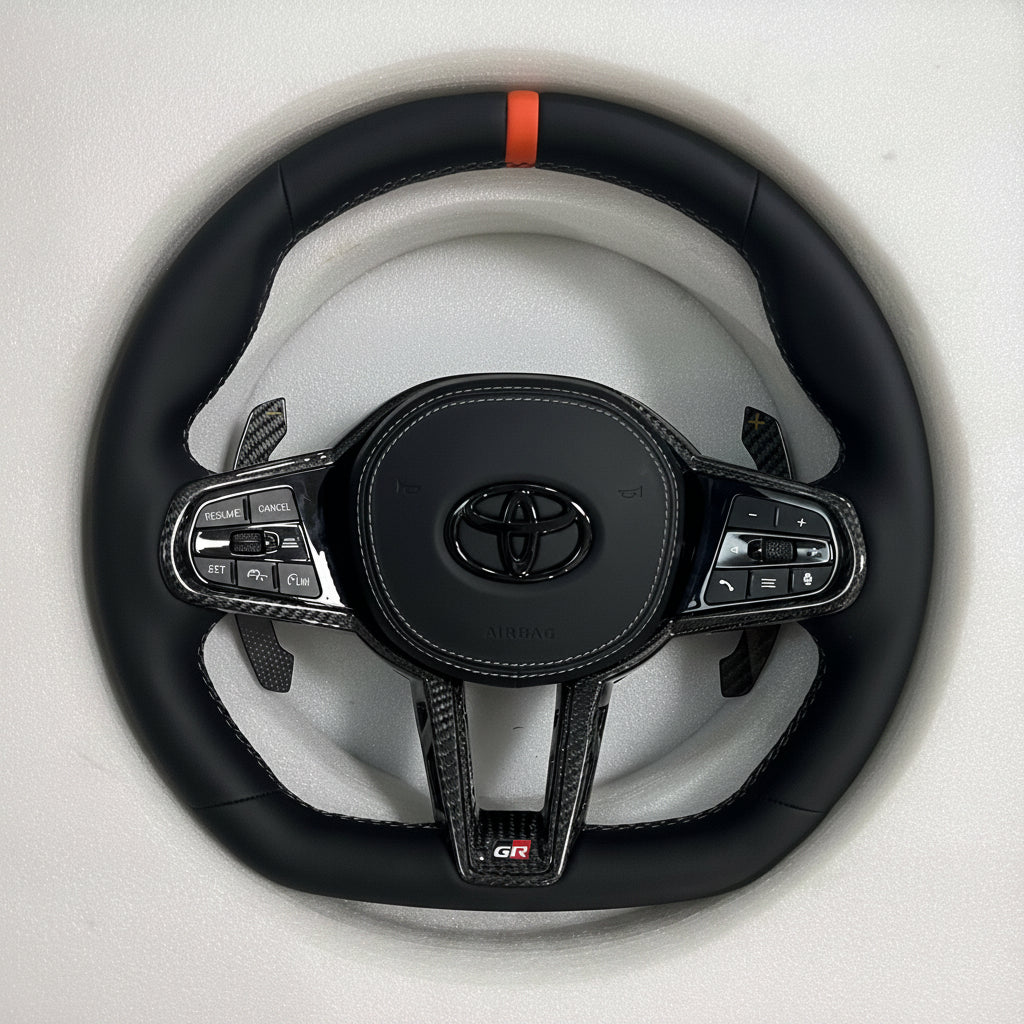 Supra steering wheel with orange center stripe, smooth leather grip, flat bottom design, carbon fiber trim, paddle shifters, and OEM airbag fitment.